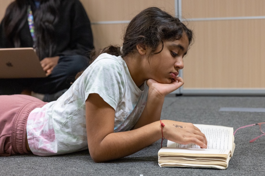 A student lies on the floor reading a chapter book, chin in hand.