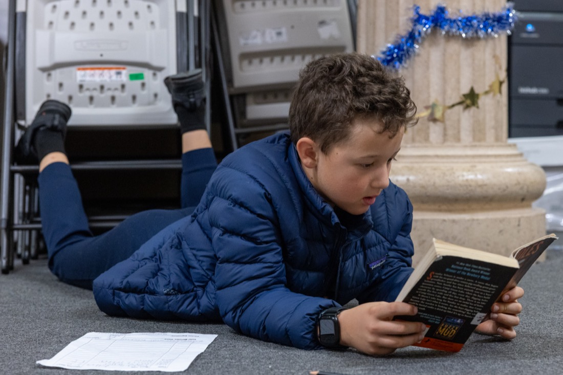 A boy reads a paperback on the floor with his reading log beside him.