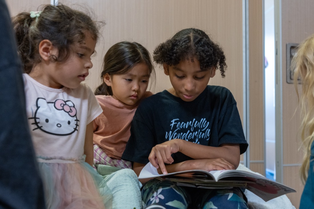 A middle schooler reads to two younger girls leaning in close.