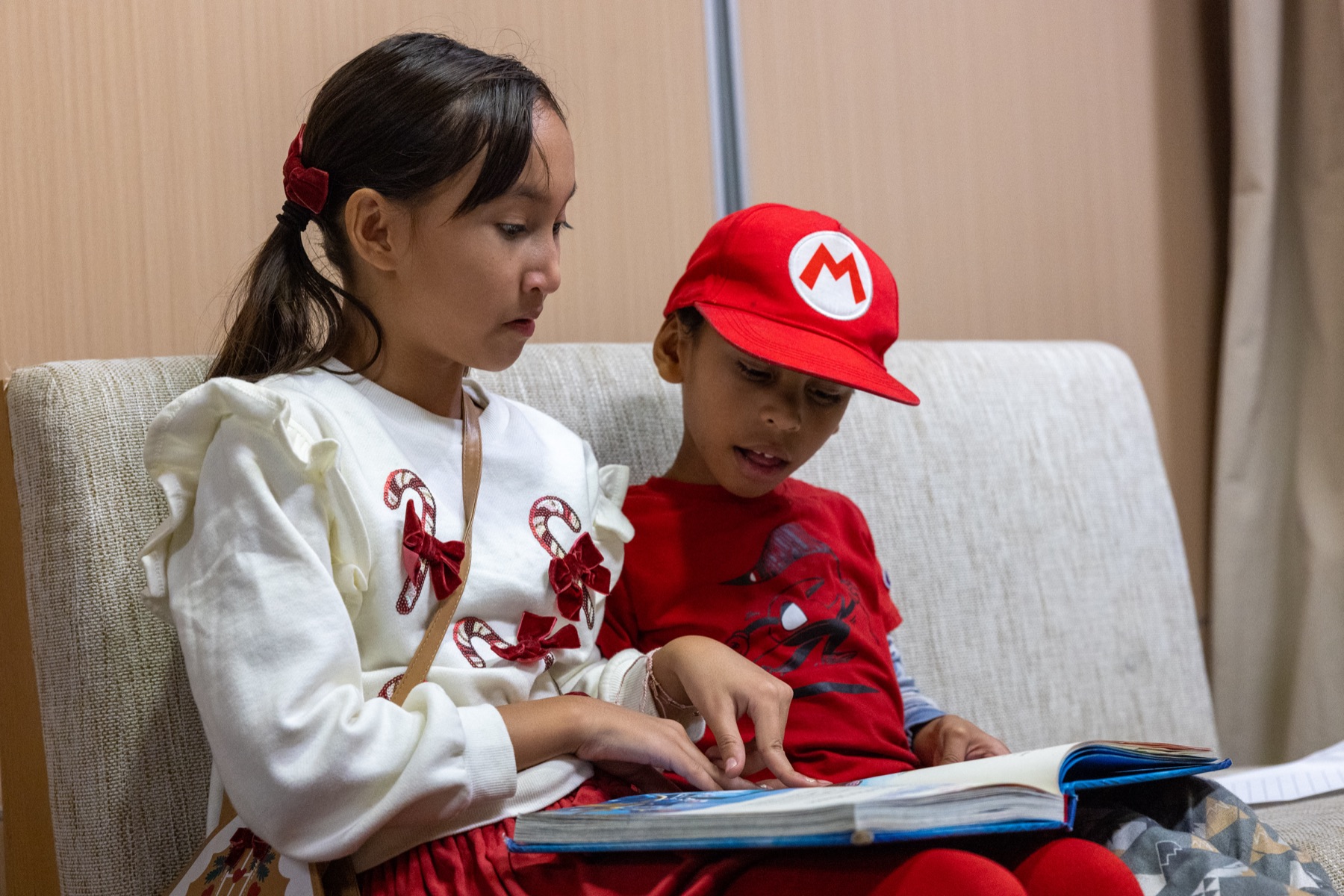 An ASTEME student points to the page while reading a picture book with a younger boy in a red cap.